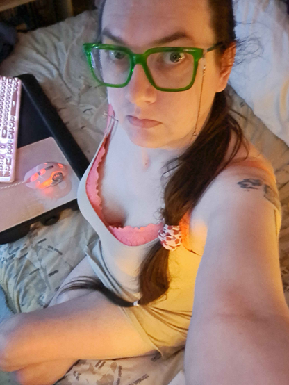 Selfie of Bunya sat on the edge of its bed, its long brown hair is tied into a ponytail with extra hair ties down the length of it and draped over its shoulder.

It is wearing a tight, beige coloured, vest top that is long enough to act as a dress, over a baby pink pushup bra that make its tits look quite nice actually, and its large framed bright green glasses with silver chains on the arms.

It is looking up at the camera with ts pale blue eyes, and its bare legs are crossed. Its Companion Cube Graffiti tattoo is just barely visible on its upper arm.