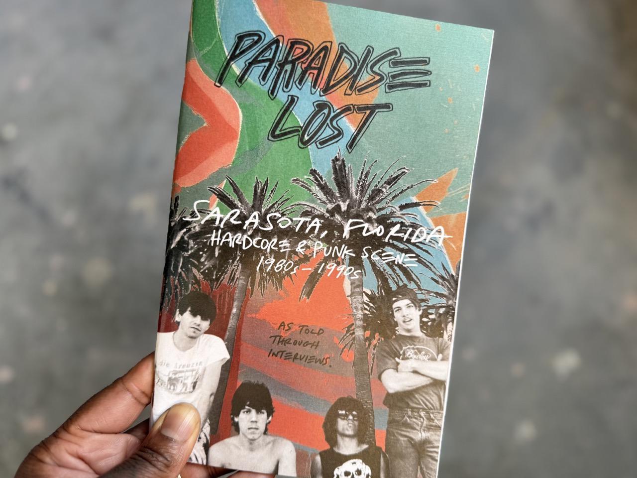Photo of my hand holding a copy of a zine. It is half-letter size and the cover is in color. The background is a colorful abstract expressionist painting by Sarasota artist Syd Solomon, using different tones of blue, yellow, pink, green and red. The title "Paradise Lost" is at the top in black sharpie, outlined. The words "Sarasota, Florida, hardcore & punk scene, 1980s-1990s" is in white handwriting on top of black & white digitally cut & pasted palm trees. A black and white cut-out photo of the Sarasota-Bradenton band BP is below. Above their heads are the handwritten words "As told through interviews."